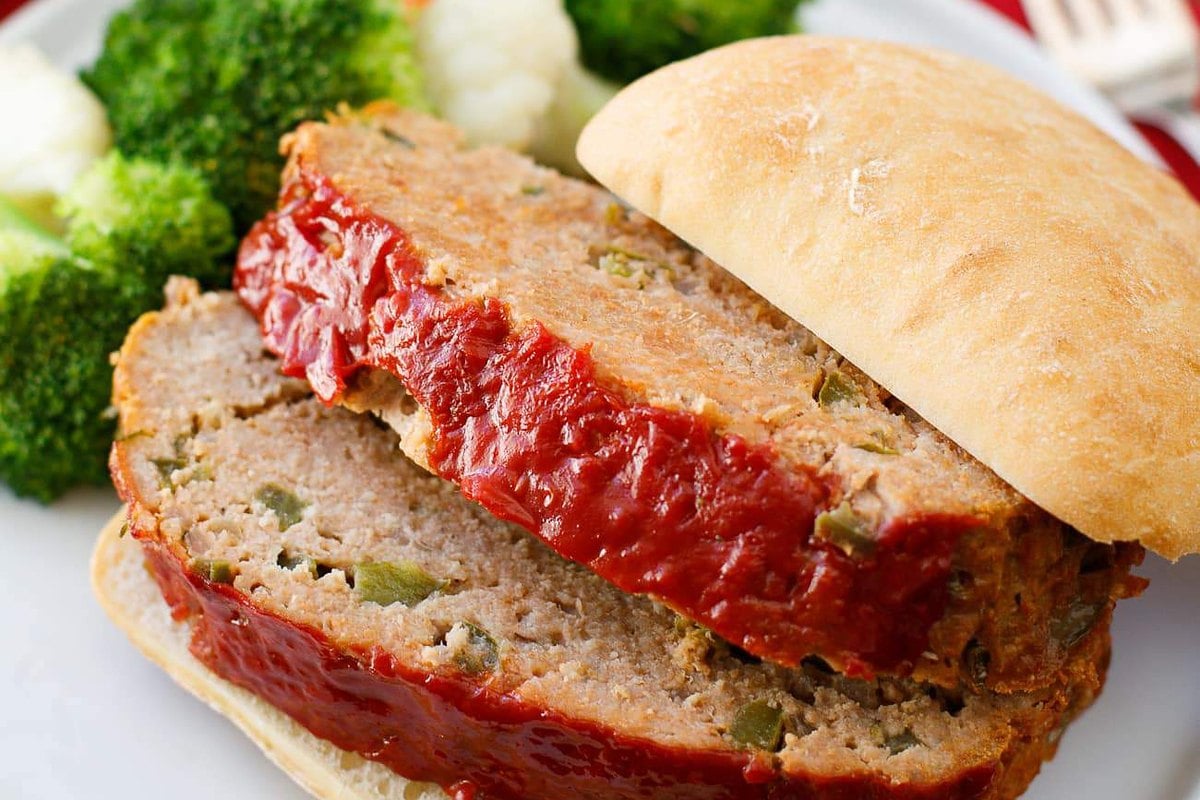 Two slices of ground turkey meatloaf with ketchup glaze on a white plate with a soft roll and broccoli