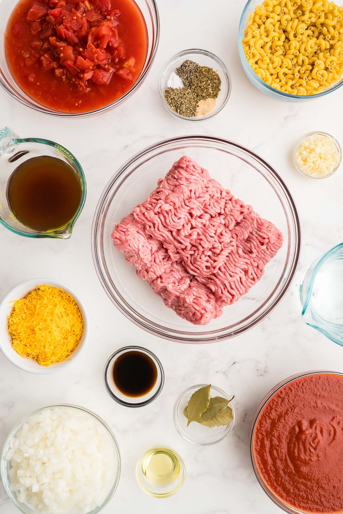 Recipe ingredients on a marble counter, including elbow macaroni, diced tomatoes, tomato sauce, beef broth, chopped onions, cheddar, and bay leaves