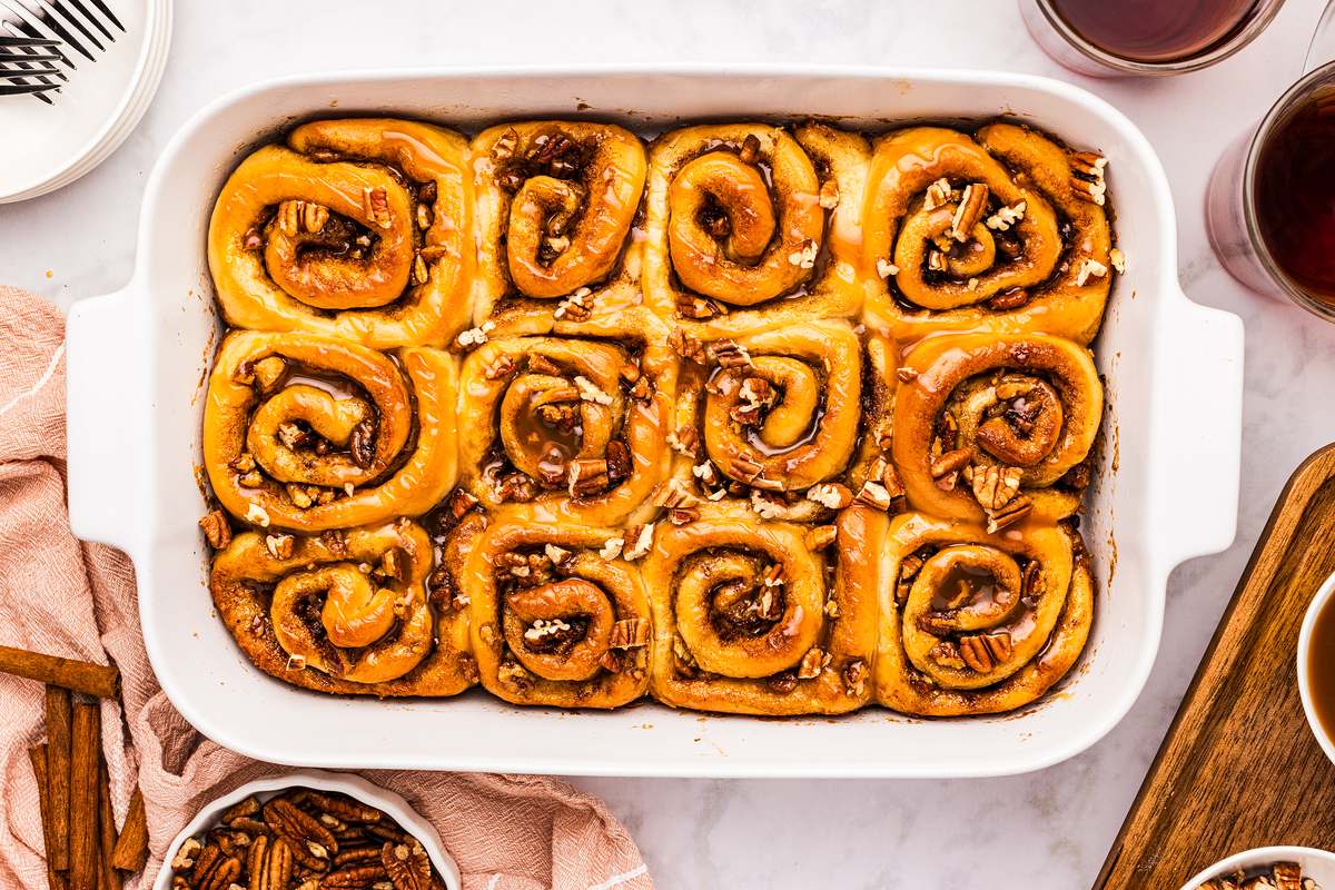 Pan of baked caramel pecan cinnamon rolls on a table with coffee and extra pecans.