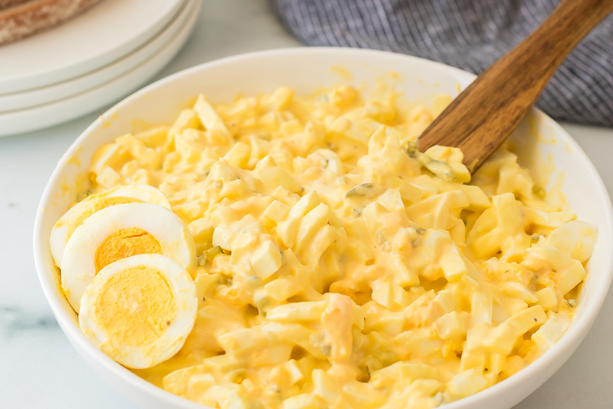 Bowl of creamy egg salad with halved hard-boiled eggs and a wooden spoon.