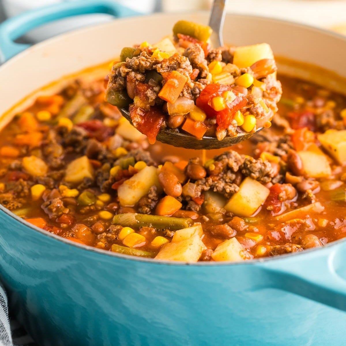 A ladle lifting hearty soup with ground beef, potatoes and corn from a pot