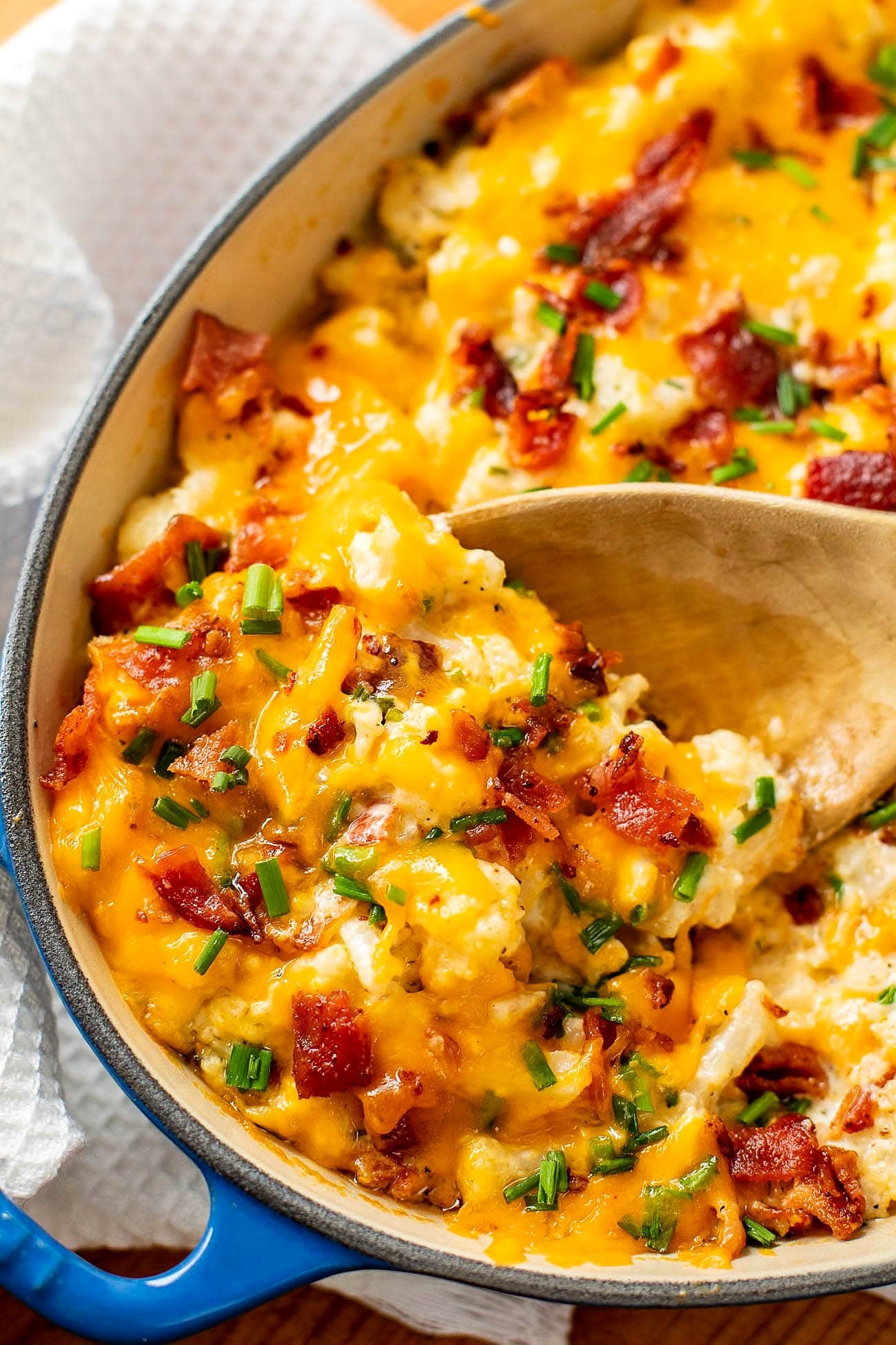 A wooden spoon scooping creamy cauliflower, bacon and melted sharp cheddar from the dish.