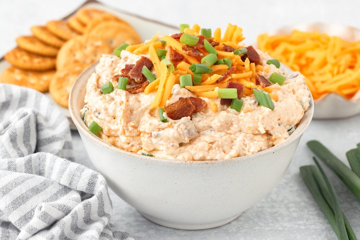 Creamy bacon and cheddar appetizer in a white bowl with crackers and shredded cheese behind it