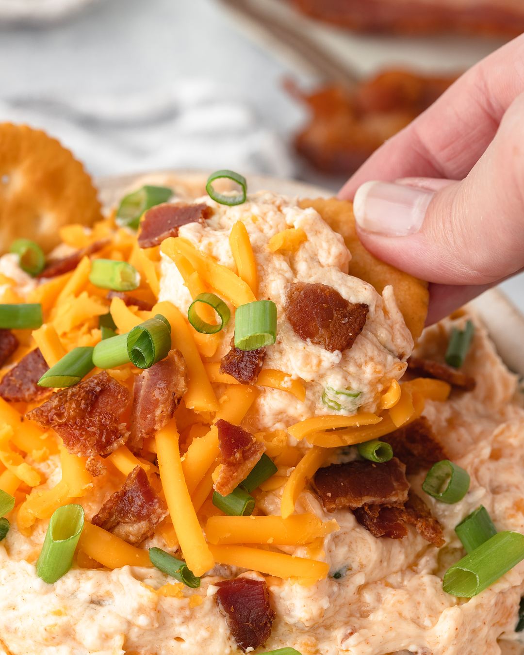 Cracker scooping up bacon, cheddar and green onions from a creamy bowl