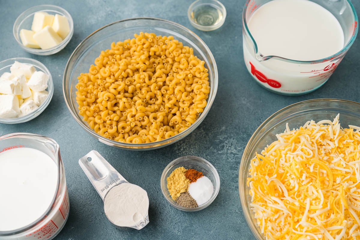 Ingredients on a blue table, including elbow pasta, shredded cheese, milk, butter, flour and seasonings.