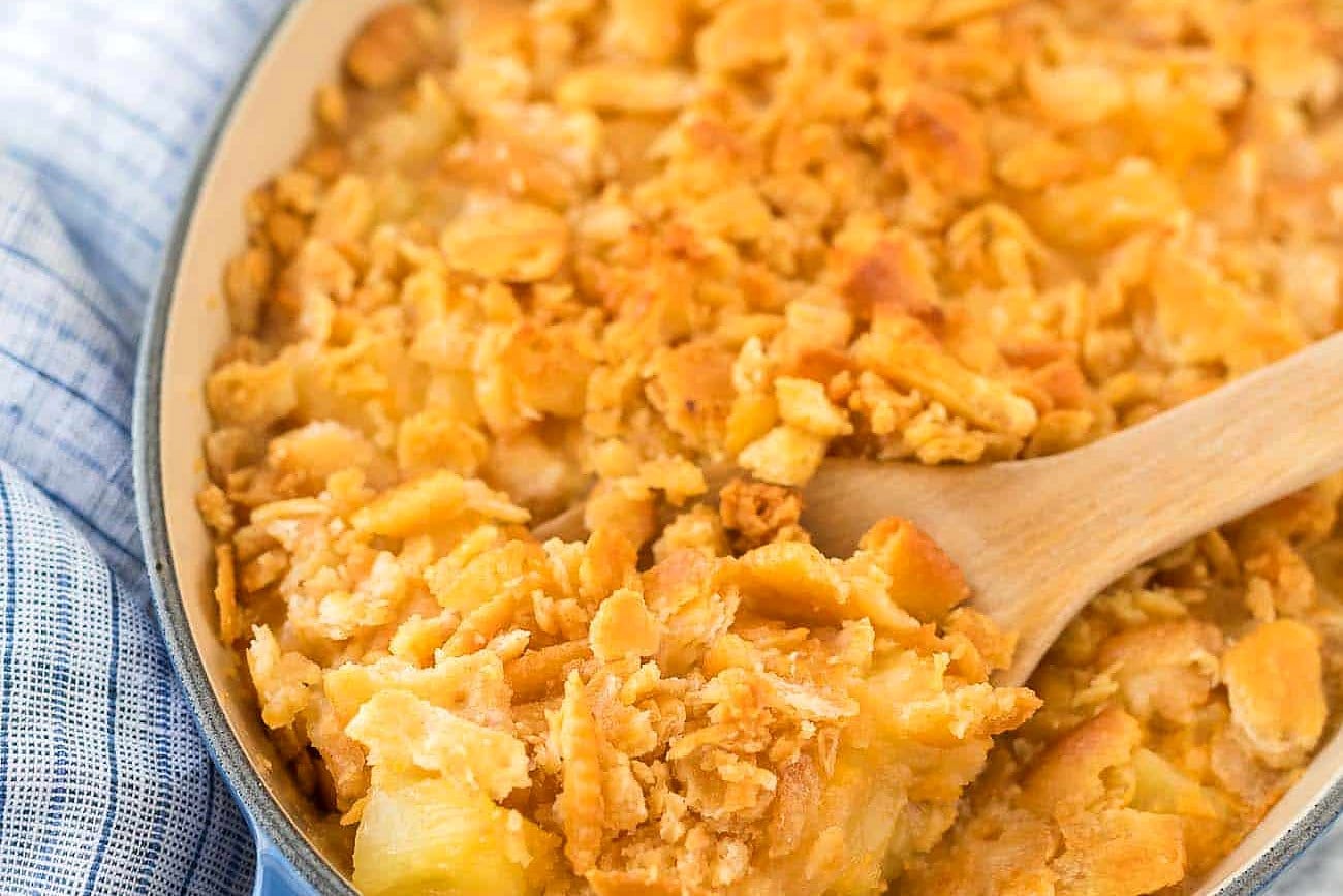 Wooden spoon scooping baked pineapple casserole with a golden Ritz cracker topping from a blue dish